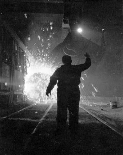 Black & white image of a silhouetted steel worker gesturing upwards as molten metal pours forth from a vat in front of him, sending glowing sparks in every direction.