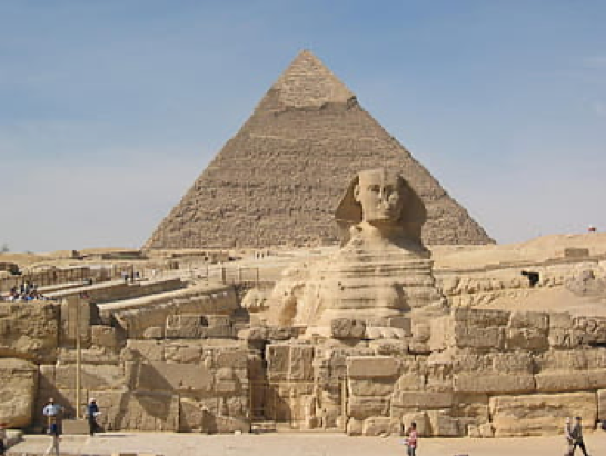  A large pyramid at Giza rises towards a hazy, white-blue sky, its grey-brown coloring mimicked by the sand, stone, and the famous sphinx in the foreground of the image. 