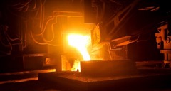 Golden light burns bright from molten metal poured out of a furnace as tubes and large industrial machines can be seen, dark compared to the furnace opening.