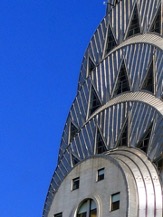 A close-up of the very top section of a skyscraper which shows a pyramid-like structure with intricate rectangular windows set into the steel frame at each different section of the tapered roof and a large circular outcropping that resembles where a large clock might go that also has windows and a blue sky behind.