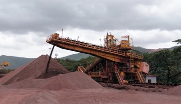 An iron mining operation shows a large, elevated conveyor belt, that resembles a wide, squat crane, pouring material to the earth where mounds of dirt reside, with a background of a forest and green hills under a cloudy sky. 