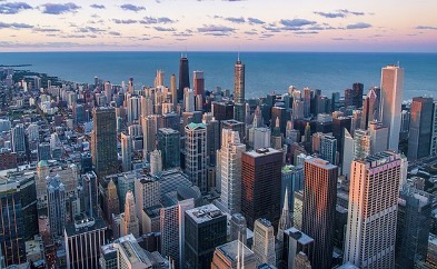 An aerial, panoramic shot of the Boston skyline filled with skyscrapers of different heights and shapes overlooking the bay, underneath a sky spotted with clouds.