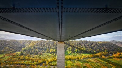 A picture of the underside of a steel bridge spanning a valley full of green trees, fields, and pastures.