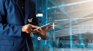 Angle on a man in a blue blazer standing in a steel shipping facility typing on his automated transaction processing device. A graphic design overlays the image, consisting of various icons representing Steel Supply Chain Management.