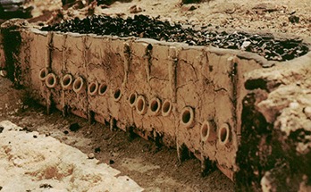 A flat, browned, dry dirt surface that looks like a cliff or box with various round orifices and cracks along the front wall and black rocks on top.