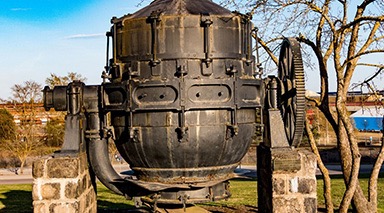 A photo of a black Bessemer Converter standing on top of two high panels made of bricks taken during a cloudless day. Behind it are leafless trees and buildings.