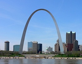 Behind a river and a parking lot full of cars a large silver arch rises precipitously towards the sky, and behind the arch can be seen a capitol building with Roman columns and a green domed roof, around which are various other tall buildings of different shapes and heights.