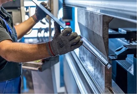 A gloved metalworker wearing heavy protective gloves and overalls feeds a steel plate into a machine which bends the steel into an “L” shape at the tip.