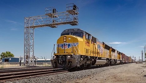 A yellow locomotive pulls a long line of containers down a train track bordered by gravel and dirt in an industrial area under a mostly clear blue sky.