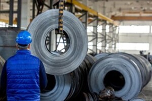 A person wearing a blue helmet and jacket is observing large rolls of steel or some other metal material stored in an industrial setting. The picture shows an industrial environment with multiple machines and equipment producing or processing metal.