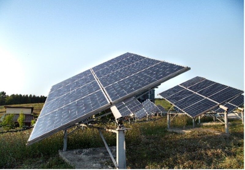 Photovoltaic solar power station with multiple panels outdoors. The panels are tilted at an angle to capture sunlight efficiently. They are mounted on metal structures and surrounded by grassy terrain. In the background, there is a clear sky and some trees.