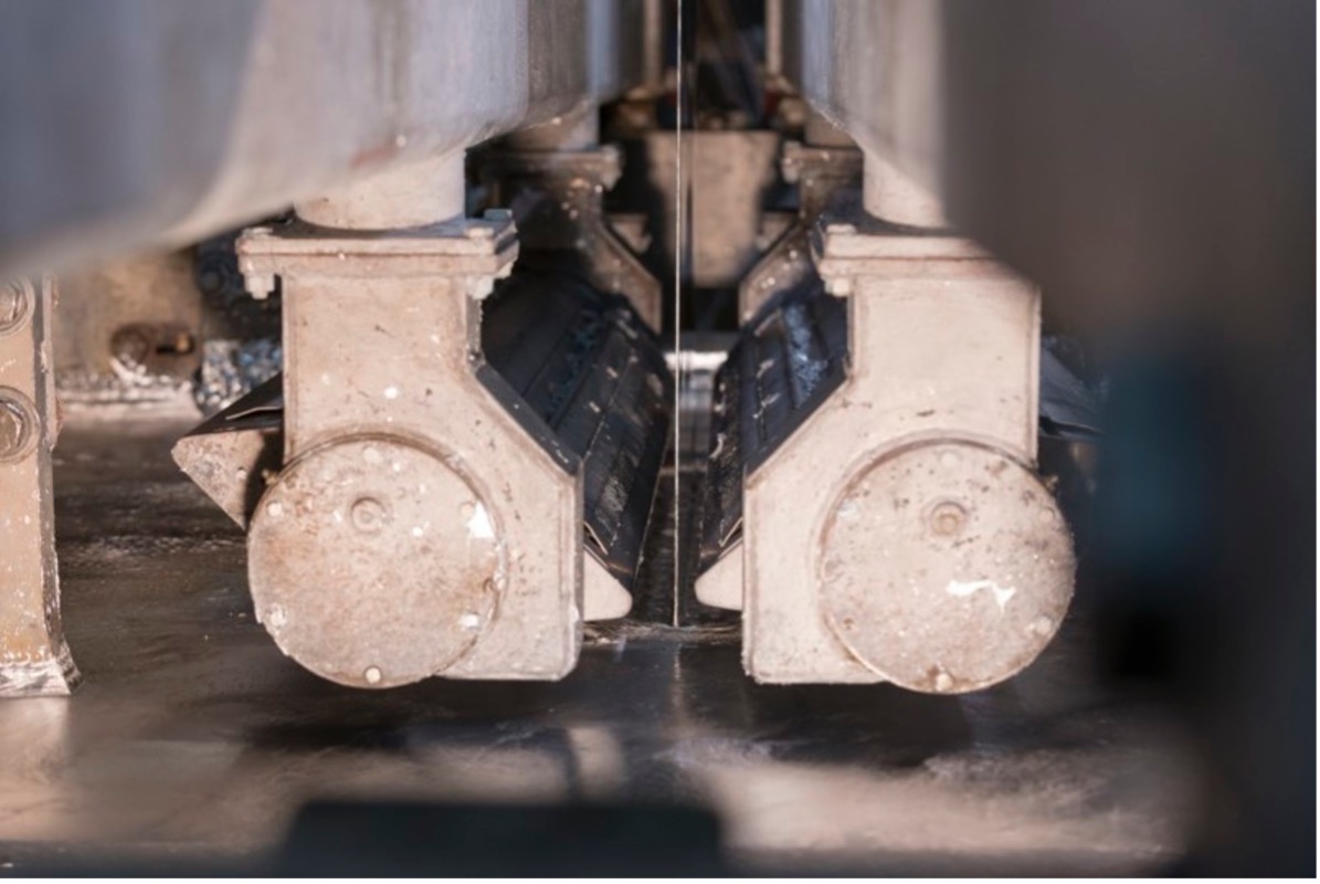 A detailed view of machinery focusing on two robust metallic wheels connected to a track or conveyor belt system. The wheels exhibit signs of wear, indicating usage, while the track consists of metal plates precisely linked together. Shadows cast by the components add depth to the image, emphasizing the mechanical intricacies.