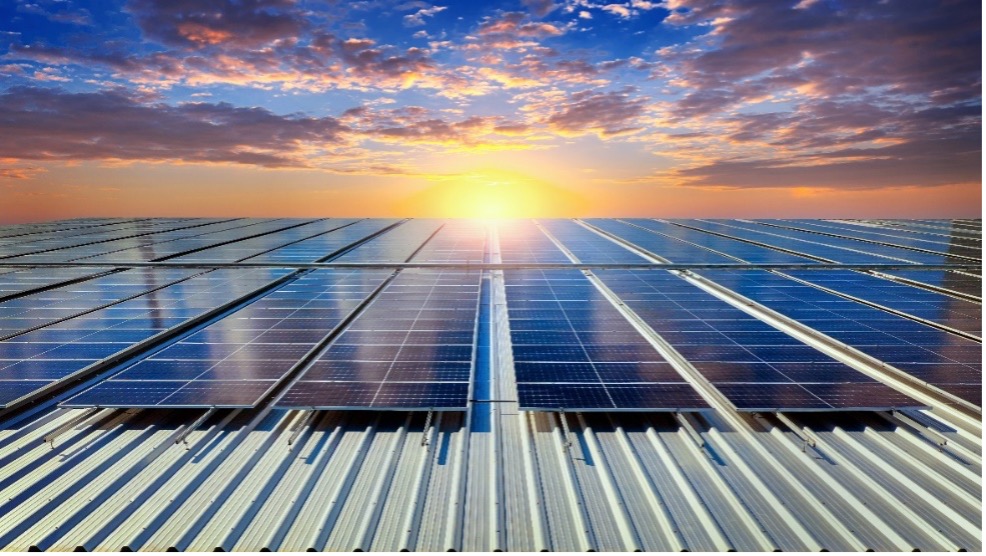 A vast array of solar panels is captured at sunset, where the sky is painted with orange, blue, and purple hues as the sun descends on the horizon. The foreground prominently features numerous solar panels installed in parallel rows.