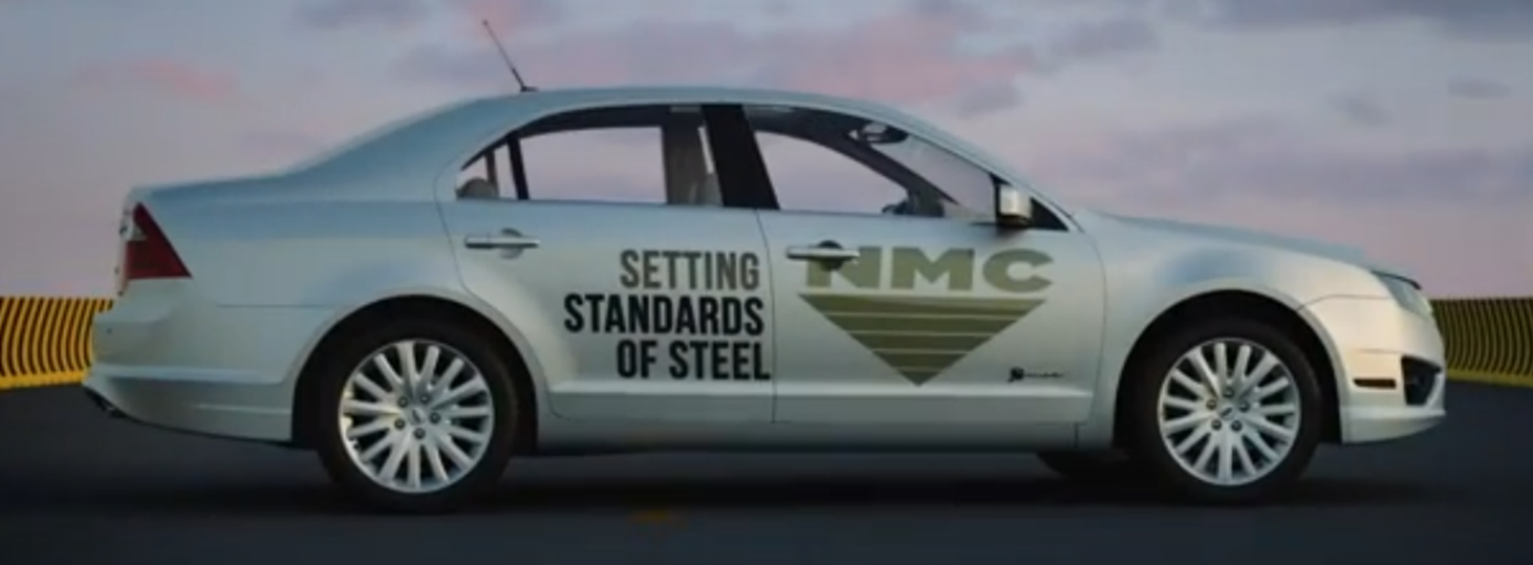 A silver four-door sedan with the text “SETTING STANDARDS OF STEEL” and “NMC” on its passenger side doors rests on the surface of a road or track. A yellow barrier is visible in the background. The sky behind the car displays hues of blue and pink.