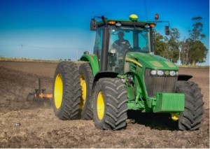 A modern sturdy green tractor, with the NMC logo overlaid, working in a tilled field.