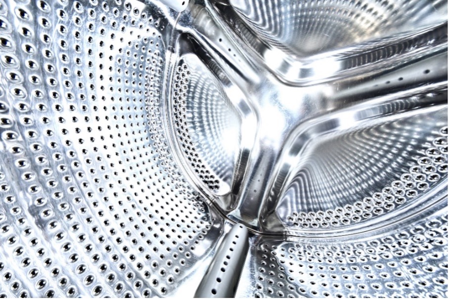 The shiny interior of a clothes washing machine. The central focus is on the inner drum, which is perforated with numerous small holes to allow water to flow in and out during the wash cycle. The drum’s metallic surface is clean and reflective, showing the intricate design that helps agitate and clean clothes.