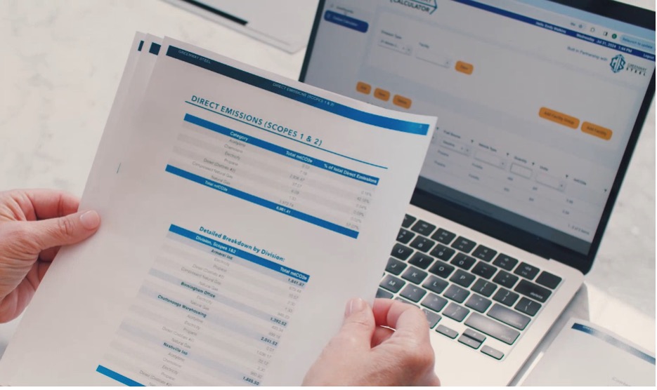 Hands holding a report on direct emissions for scopes 1 and 2, with a laptop in the background displaying a carbon footprint calculator interface